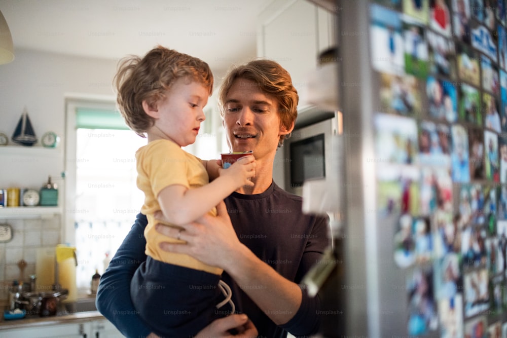 Um pai feliz com um filho pequeno dentro de casa na cozinha de casa, abrindo geladeira.
