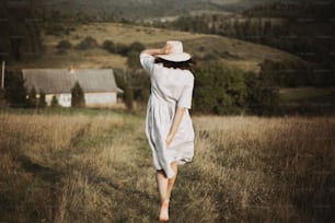 Stylish girl in linen dress and hat running barefoot in grass in sunny field at village. Boho woman relaxing in countryside, simple rustic life. Atmospheric image. Space text