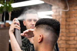 Peluquero profesional que usa laca para el cabello en el cabello del cliente en la peluquería, el peluquero fresco salpica de la botella de spray en el cabello de su cliente.