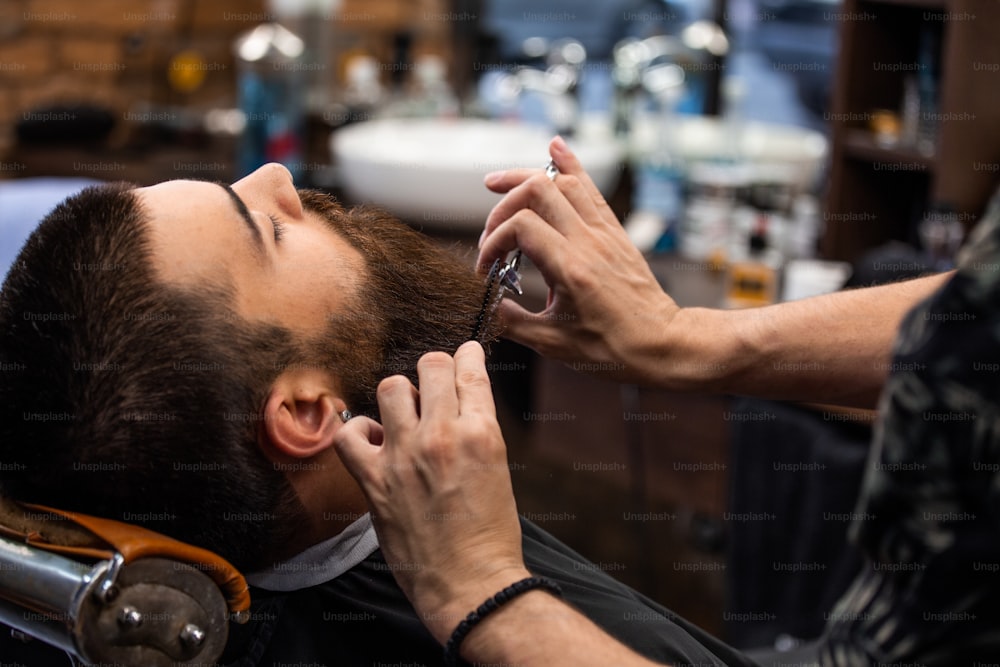 Nahaufnahme eines zugeschnittenen Fotos der Arbeit eines Friseurs für einen attraktiven Mann im Friseursalon. Er stylt seinen Bart mit einer Schere