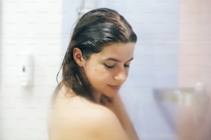 Joven mujer feliz duchándose en casa o en el baño del hotel. Hermosa chica morena lavándose el pelo y disfrutando de un tiempo de relax. Higiene corporal y de la piel, concepto de estilo de vida
