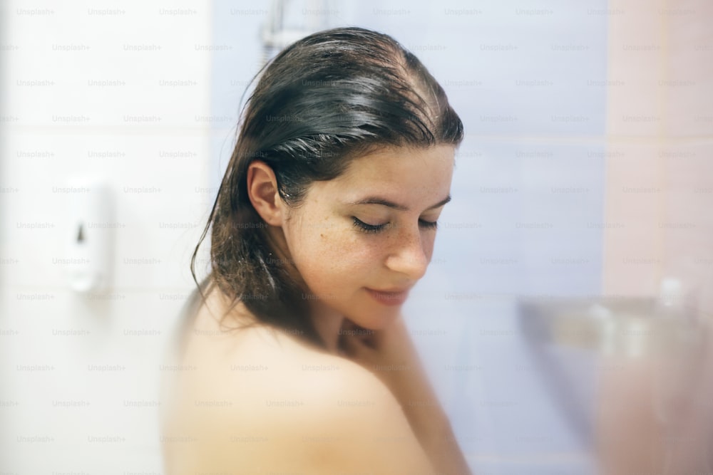 Young happy woman taking shower at home or hotel bathroom. Beautiful  brunette girl washing her hair and enjoying relax time. Body and skin hygiene, lifestyle concept