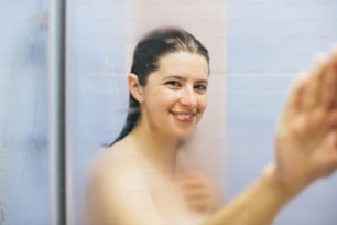 Young happy woman taking hot shower at home or hotel bathroom. Beautiful brunette girl holding hand on glass with steam, enjoying time in shower. Body and skin hygiene, lifestyle concept