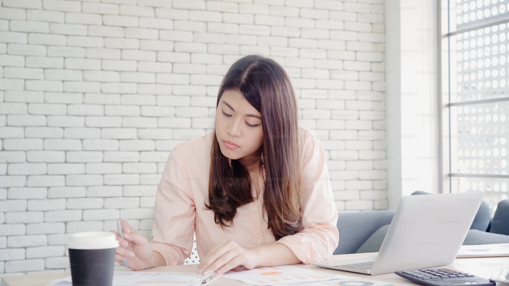 Beautiful young smiling asian woman working laptop on desk in living room at home. Asia business woman writing notebook document finance and calculator in home office. Enjoying time at home concept.