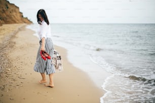Femme bohème heureuse se relaxant en mer, profitant d’une promenade sur une île tropicale. Fille hipster élégante marchant pieds nus et souriant sur la plage, tenant un sac et des chaussures à la main. Vacances. Espace pour le texte