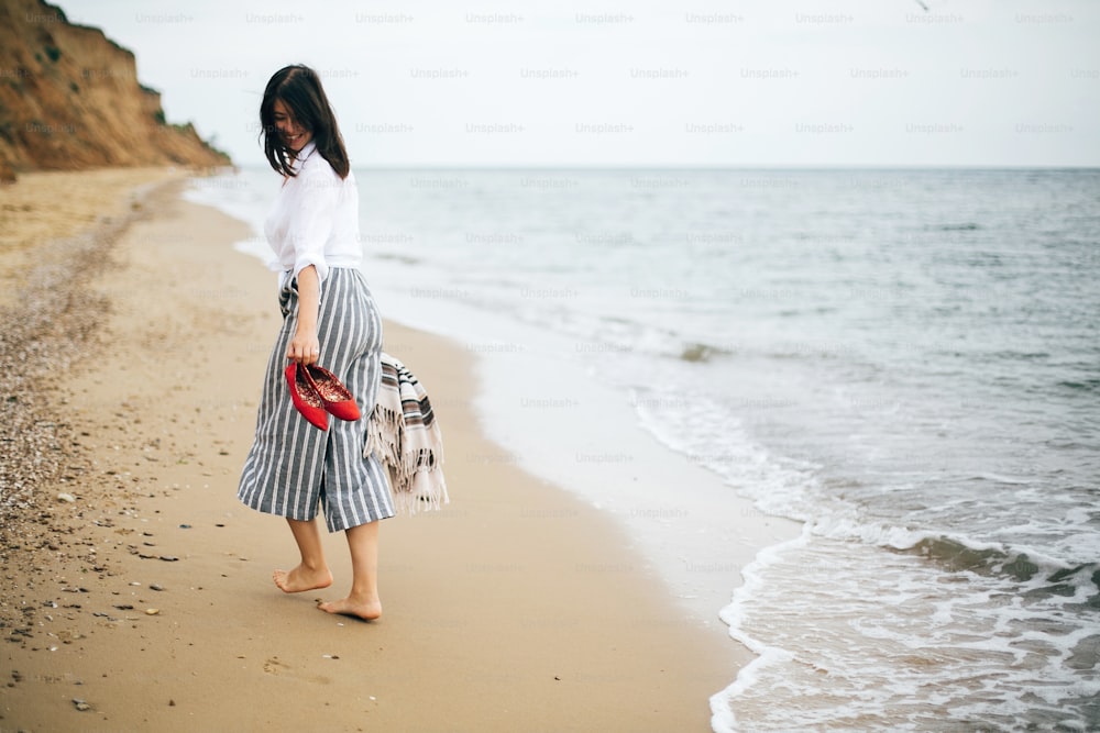 Glückliche Boho-Frau, die sich auf See entspannt und einen Spaziergang auf einer tropischen Insel genießt. Stilvolles Hipster-Mädchen, das barfuß und lächelnd am Strand läuft, Tasche und Schuhe in der Hand hält. Sommerferien. Platz für Text