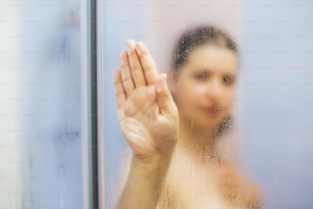 Young happy woman taking hot shower at home or hotel bathroom. Beautiful brunette girl holding hand on glass with steam, enjoying time in shower. Body and skin hygiene