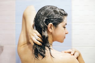 Giovane donna felice che si lava i capelli con lo shampoo, schiumando con le mani. Bella ragazza bruna che fa la doccia e si gode il tempo di relax. Igiene del corpo, dei capelli e della pelle, concetto di stile di vita.