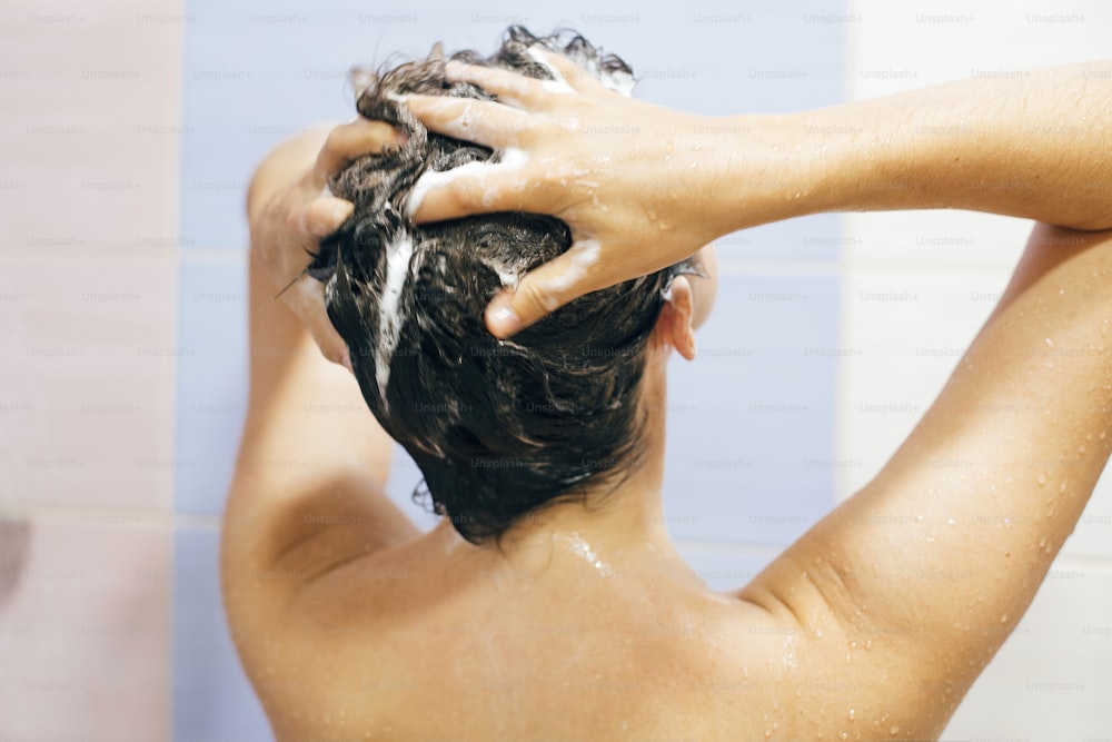 Young happy woman washing her hair with shampoo, hands with foam closeup. Back of beautiful brunette girl taking shower and enjoying relax time. Body and hair hygiene, lifestyle concept