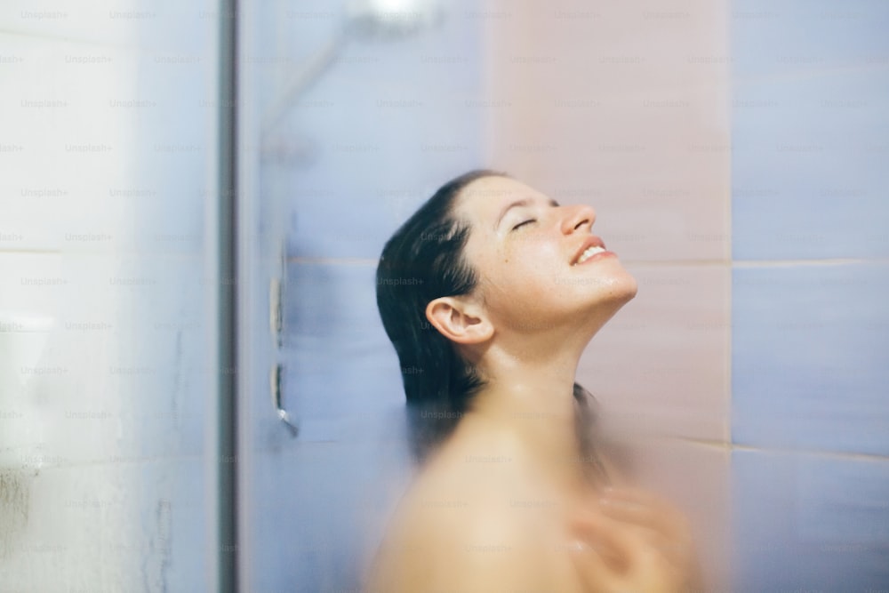 Young happy woman taking hot shower at home or hotel bathroom. Sensual portrait of beautiful brunette girl enjoying time in shower. Body and skin hygiene, lifestyle concept. Space text