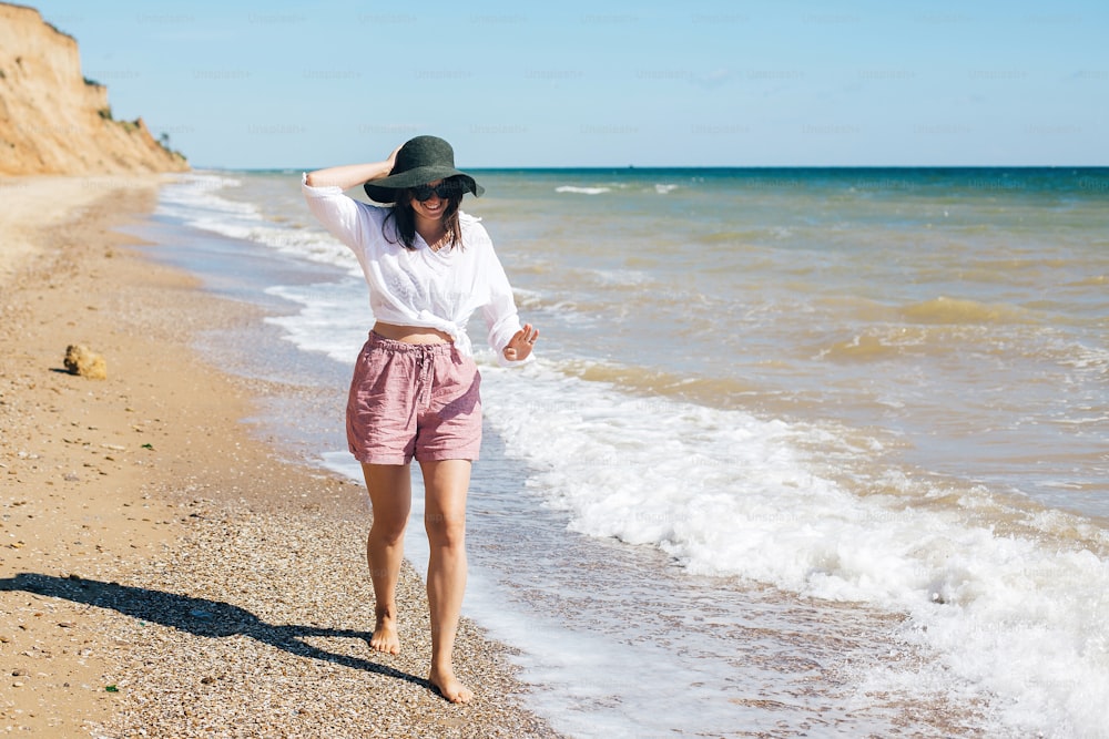 Stylish hipster girl in hat walking on beach and smiling. Summer vacation. Happy young boho woman relaxing and enjoying sunny warm day at tropical island and blue sky. Space for text
