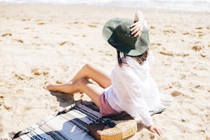 Summer vacation. Happy young boho woman relaxing and enjoying sunny warm day at ocean. Space for text. Stylish hipster girl in hat sitting on beach with straw bag and tanning