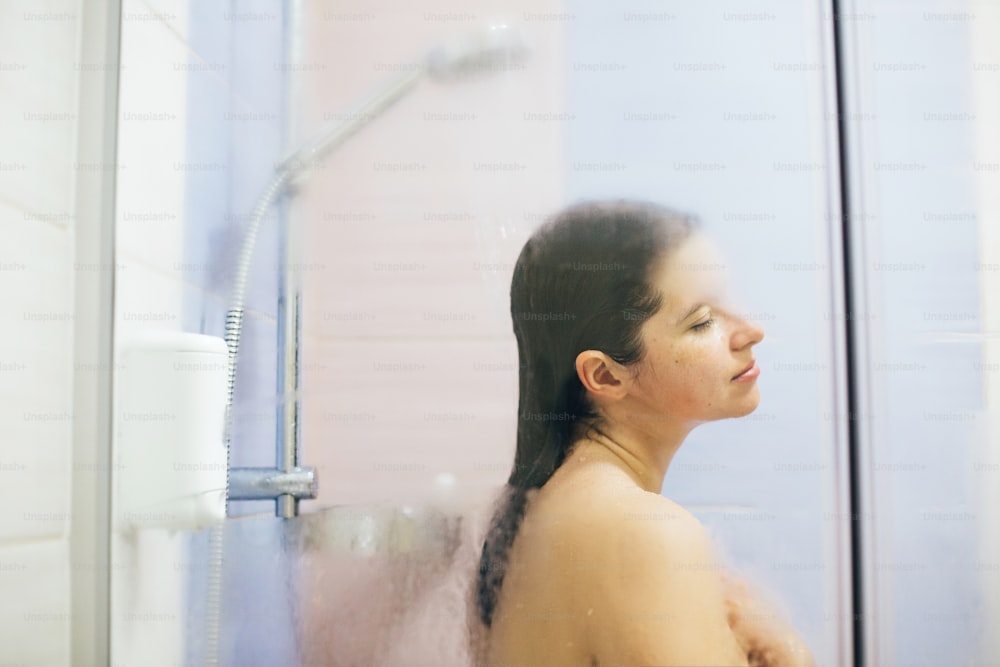 Young happy woman taking hot shower at home or hotel bathroom. Sensual portrait of beautiful brunette girl enjoying time in shower. Body and skin hygiene, lifestyle concept. Space text