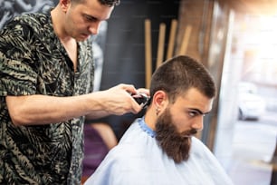 Haarschnitt perfekt aussehen lassen. Junger bärtiger Mann, der vom Friseur die Haare schneiden wird, während er auf dem Stuhl im Friseursalon sitzt