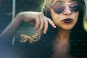 Stylish hipster girl posing in sunny street, atmospheric moment. Fashionable cool woman in black sunglasses smiling and relaxing in evening light. Selective focus. Retro effect