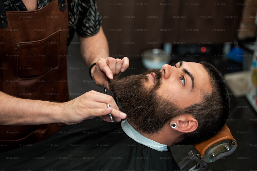 Bearded male sitting in an armchair in a barber shop while hairdresser trip his beard with