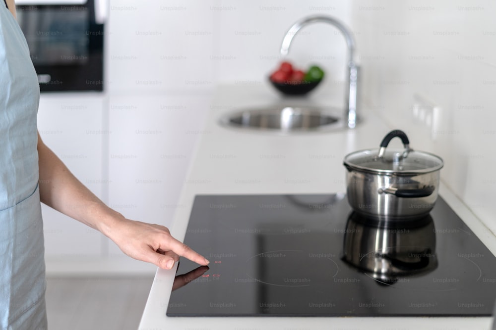 Cropped view of woman using built in stove with saucepan on top, selecting program on display for cooking, standing at white modern kitchen