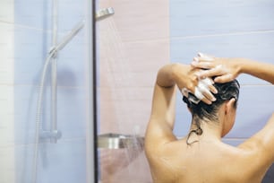 Young happy woman washing her hair with shampoo, foaming with hands. Beautiful  brunette girl taking shower and enjoying relax time. Body, hair and skin hygiene, lifestyle concept.