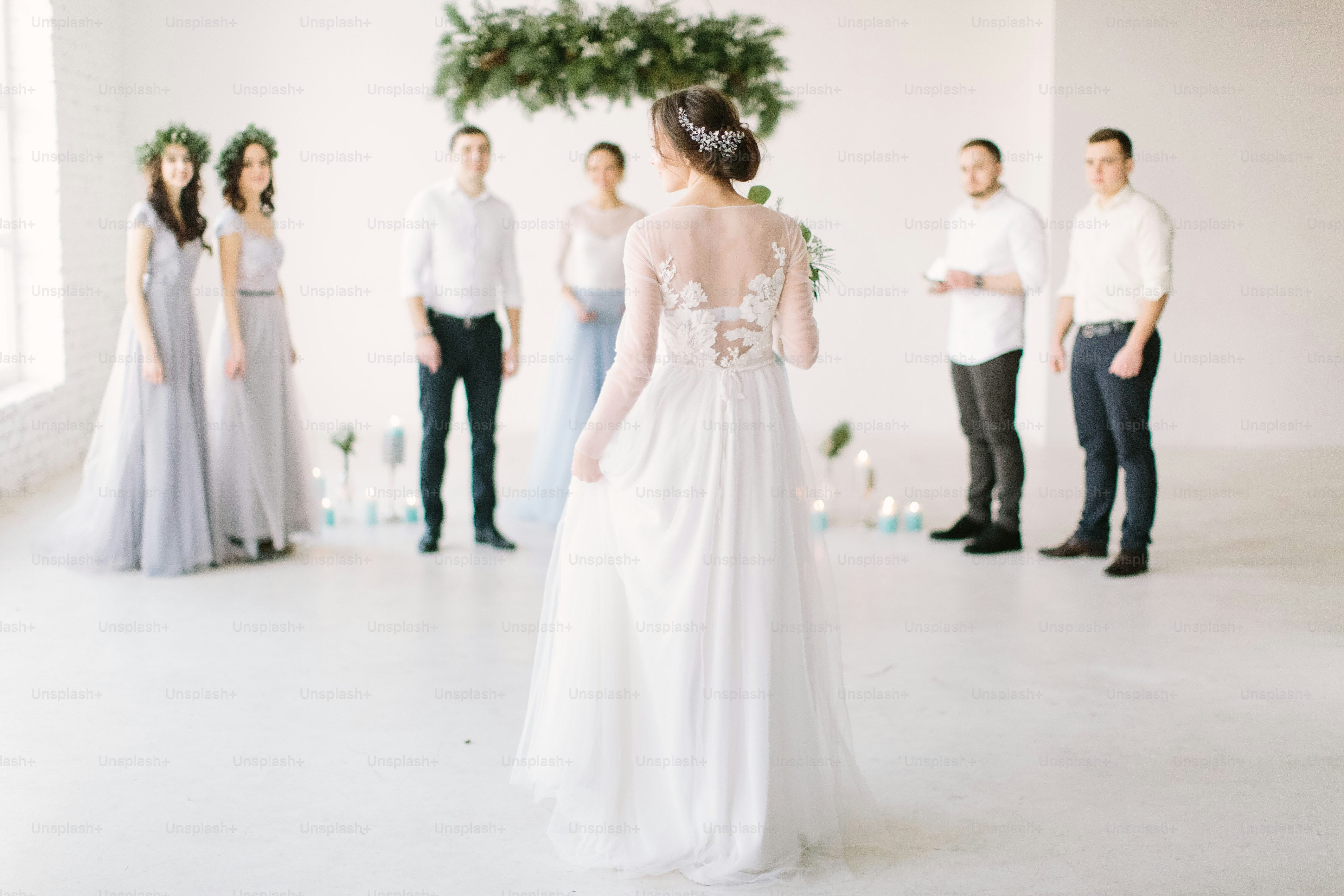 Pretty bride in a luxury white dress holds a wedding bouquet and is going to her groom for wedding ceremony. White room is decorated with pine, flowers and blue candles.