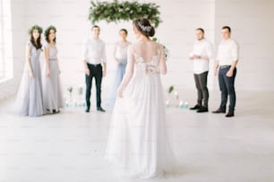 Die hübsche Braut in einem luxuriösen weißen Kleid hält einen Hochzeitsstrauß und geht zur Hochzeitszeremonie zu ihrem Bräutigam. Das weiße Zimmer ist mit Kiefernholz, Blumen und blauen Kerzen dekoriert.