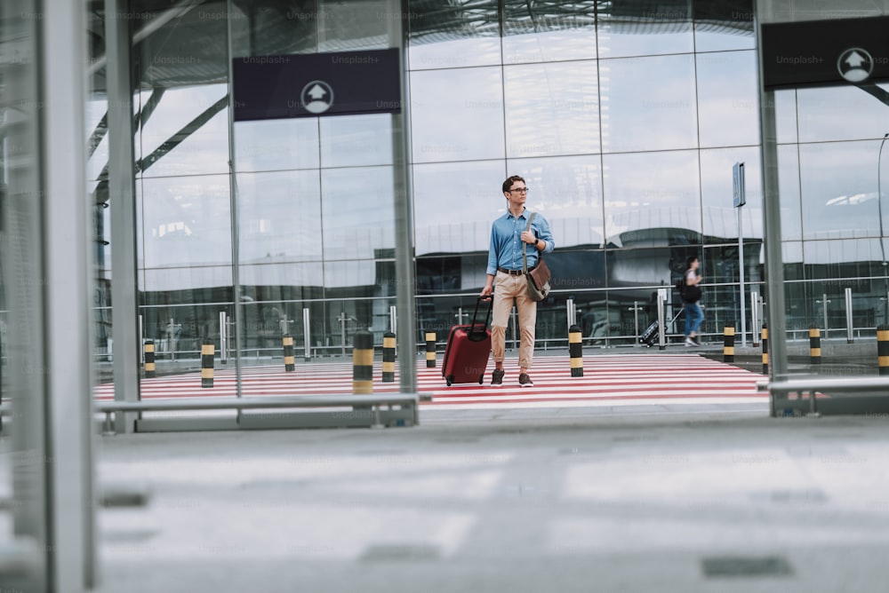 Full length portrait of man in glasses holding bag and standing near crosswalk with airport on background. Copy space in left side