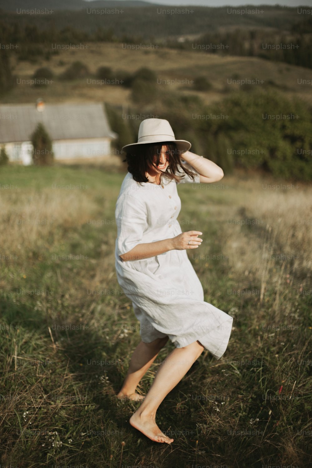 Stylish girl in linen dress running barefoot among herbs and wildflowers in sunny field in mountains. Boho woman relaxing in countryside, simple rustic life. Atmospheric image. Space text