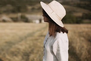 Stylish girl in linen dress and hat walking in sunny field grass at village  in mountains. Boho woman relaxing in countryside, simple rustic life. Atmospheric image. Space text