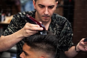 Close up shot of man getting trendy haircut at barber shop. Male hairstylist serving client, making haircut using machine and comb.