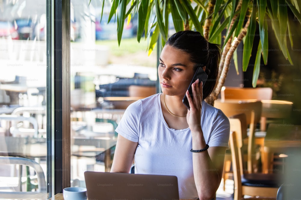 Junge Frau mit Kaffeetasse und Laptop beim Telefon. Frau macht eine Pause. Geschäfte machen Vom Café aus
