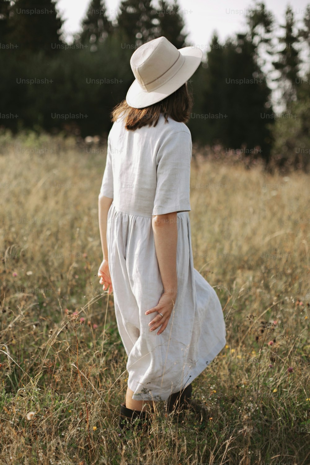 Stilvolles Mädchen in Leinenkleid und Hut, das zwischen Kräutern und Wildblumen spazieren geht und auf das Feld schaut. Boho-Frau, die sich auf dem Land entspannt, einfacher langsamer Lebensstil.  Atmosphärisches Bild