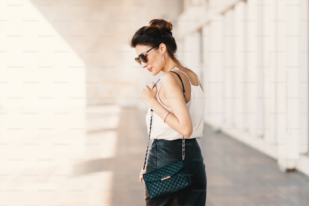 Beautiful smiling young woman in sunglasses wearing leather clutch and skirt, white easy blouse and looking away over the shoulder while walking outdoors. Spirit of the city.