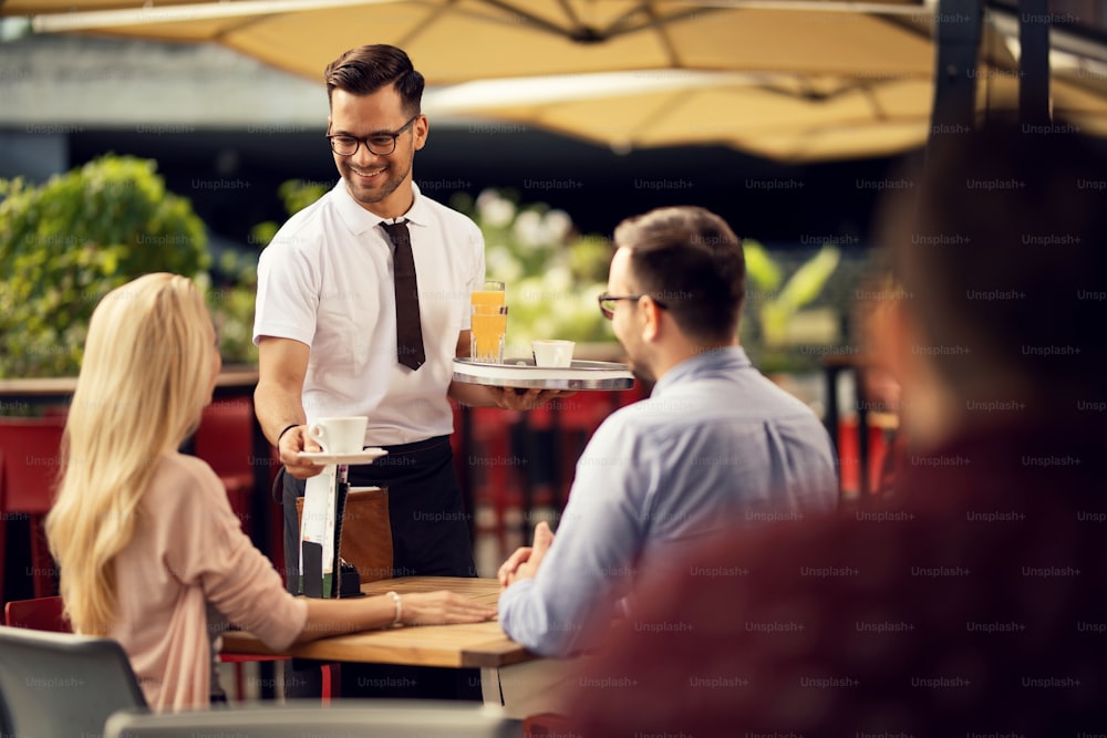 Camarero feliz dando café a una pareja mientras los sirve en una cafetería al aire libre.