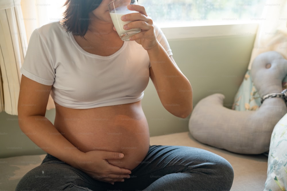 Happy pregnant woman drinks milk in glass at home while taking care of her child. The young expecting mother holding baby in pregnant belly. Calcium food nutrition for strong bones of pregnancy.