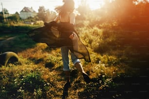 Stylish hipster girl having fun in sunny park in amazing sunbeams, atmospheric moment. Fashionable cool woman dancing in evening light, back view. Selective focus. Retro effect. Creative image