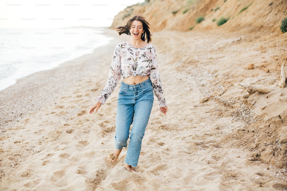 Stylish hipster girl running on beach at sea and smiling. Happy boho woman in denim jeans and floral blouse relaxing at sandy cliff on tropical island. Travel and summer vacation concept