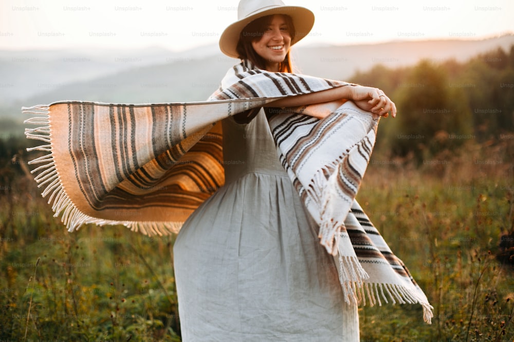 Happy hipster woman in poncho and hat enjoying traveling in evening mountains. Stylish boho girl walking in sunny light at atmospheric sunset in meadow. Space for text