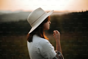 Stylish boho girl portrait in sunny light at atmospheric sunset in meadow. Calm hipster woman in poncho and hat enjoying traveling in evening mountains. Space for text