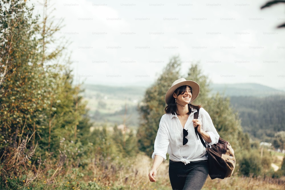 Chica hipster feliz con mochila que viaja en la cima de una montaña soleada, caminando por colinas con bosques. Mujer elegante sonriendo y disfrutando de la caminata en las montañas. Pasión por los viajes y viajes.