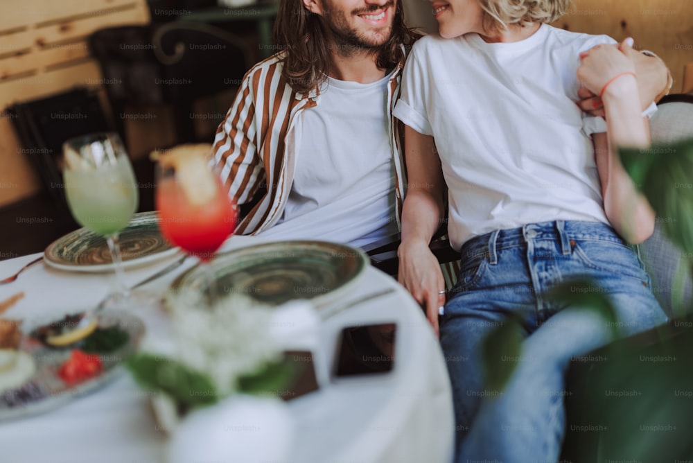 Cropped picture of bearded man wrapping arm around shoulders of his charming girlfriend while she smiling. They sitting at the table with plates and cocktails