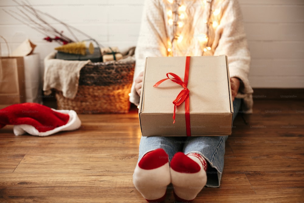 Stylish happy girl holding craft christmas gift box with red ribbon on legs in christmas lights. Young hipster woman in cozy sweater showing christmas present. Merry Christmas.