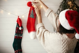 Stylish girl in cozy sweater and santa hat decorating room for christmas holidays with stockings, garland light and christmas tree on white wall . Merry Christmas. Happy Holidays