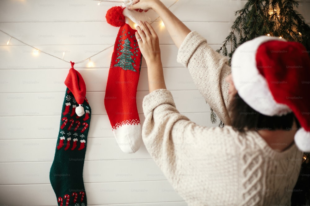 Ragazza elegante in maglione accogliente e cappello da babbo natale che decora la stanza per le vacanze di natale con calze, ghirlanda di luce e albero di natale su parete bianca. Buon Natale. Buone Feste