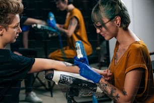 Close up tattoo artist demonstrates the process of getting black tattoo with paint. Master works in blue sterile gloves. Master of tattoo fill circuit tattoo.