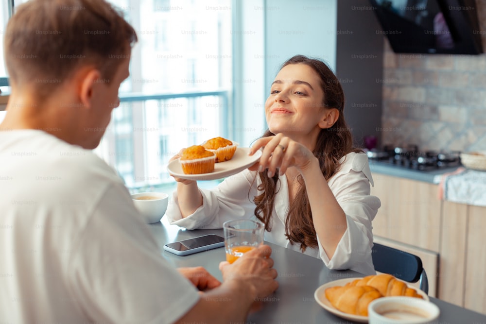 Muffins pour le mari. Femme aimante et attentionnée souriant tout en donnant des muffins pour le mari le matin