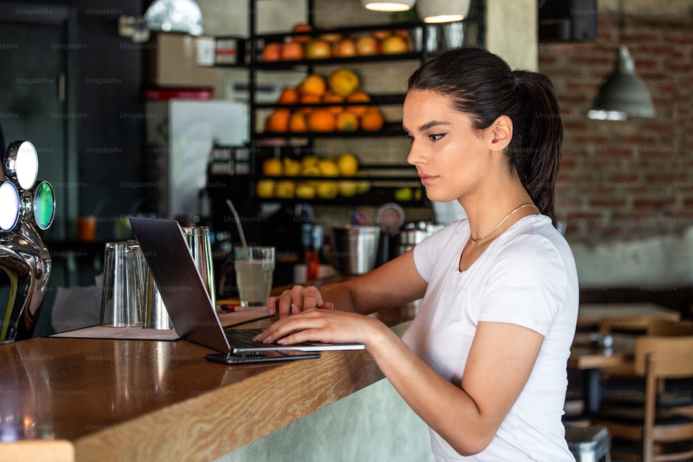 Jeune femme avec un sourire mignon assis avec un net-book portable dans l’intérieur d’un café moderne pendant le temps de loisirs, charmante étudiante heureuse utilisant un ordinateur portable pour se préparer au travail de cours