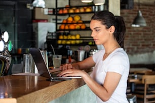 Giovane femmina con sorriso carino seduta con netbook portatile in moderno interno di caffetteria durante il tempo di ricreazione, affascinante studentessa felice che utilizza il computer portatile per prepararsi al lavoro del corso