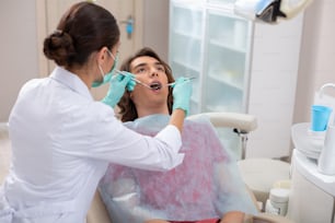 Examen de aparatos dentales. Paciente masculino con la boca abierta sentado en una silla mientras la dentista revisa sus brackets dentales