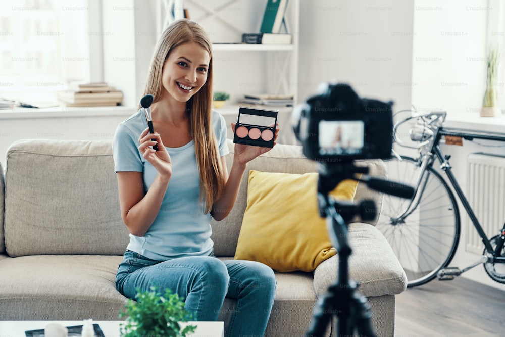 Mulher jovem bonita sorrindo e aplicando palete de maquiagem enquanto faz vídeo de mídia social em casa