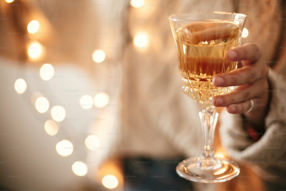 Stylish girl in cozy sweater holding champagne glass on background of christmas lights, celebrating and toasting in festive room. Space for text. Happy New Year eve party. Happy Holidays