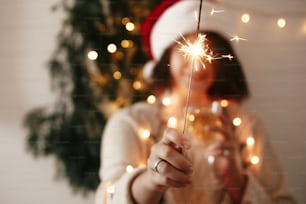 Feliz ambiente de fiesta de Nochevieja. Bengala encendida en la mano de una chica elegante en sombrero de Papá Noel en el fondo de la luz moderna del árbol de Navidad en la habitación oscura. Mujer con fuegos artificiales. Felices Fiestas